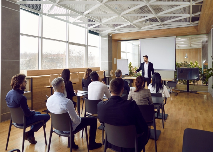 Team of people having class with business trainer. Group of male and female employees sitting at desks in modern office and listening to lecture by experienced teacher sharing knowledge and guidance